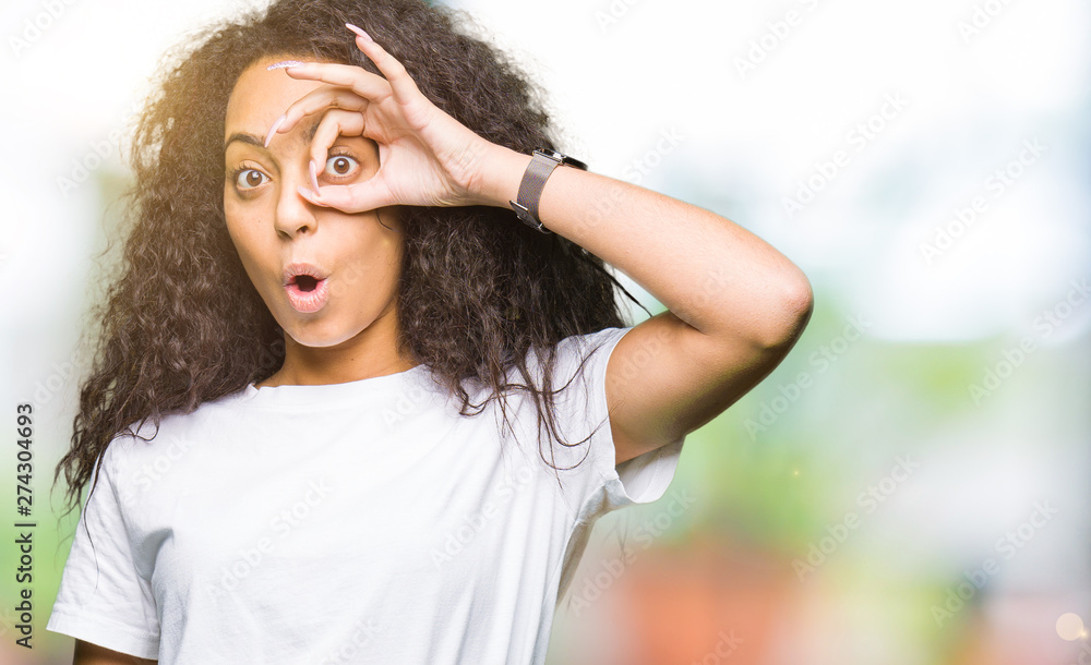 Young beautiful girl with curly hair wearing casual white t-shirt doing ok gesture shocked with surprised face, eye looking through fingers. Unbelieving expression. Young beautiful girl with curly hair wearing casual white t-shirt doing ok gesture shocked with surprised face, eye looking through fingers. Unbelieving expression.