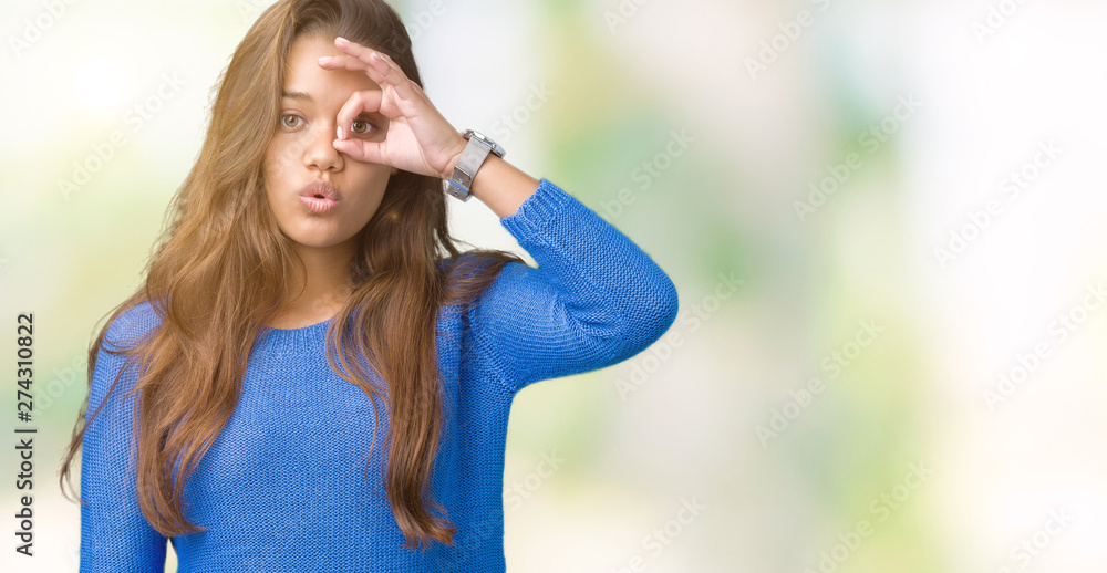 Young beautiful brunette woman wearing blue sweater over isolated background doing ok gesture shocked with surprised face, eye looking through fingers. Unbelieving expression.