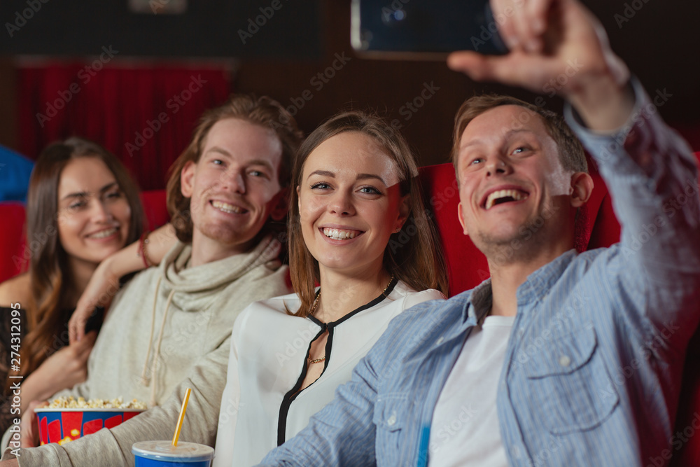 Selective focus of good looking girl looking at camera and smiling while boyfriend keeping phone and taking selfie in cinema. Cheerful company enjoying premiere and eating popcorn. Concept of movie. Selective focus of good looking girl looking at camera and smiling while boyfriend keeping phone and taking selfie in cinema. Cheerful company enjoying premiere and eating popcorn. Concept of movie.
