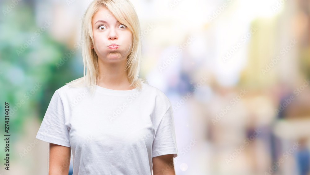 Young beautiful blonde woman wearing white t-shirt over isolated background puffing cheeks with funny face. Mouth inflated with air, crazy expression.