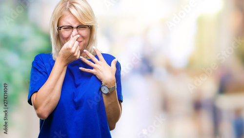 Young beautiful blonde woman wearing glasses over isolated background smelling something stinky and disgusting, intolerable smell, holding breath with fingers on nose. Bad smells concept.