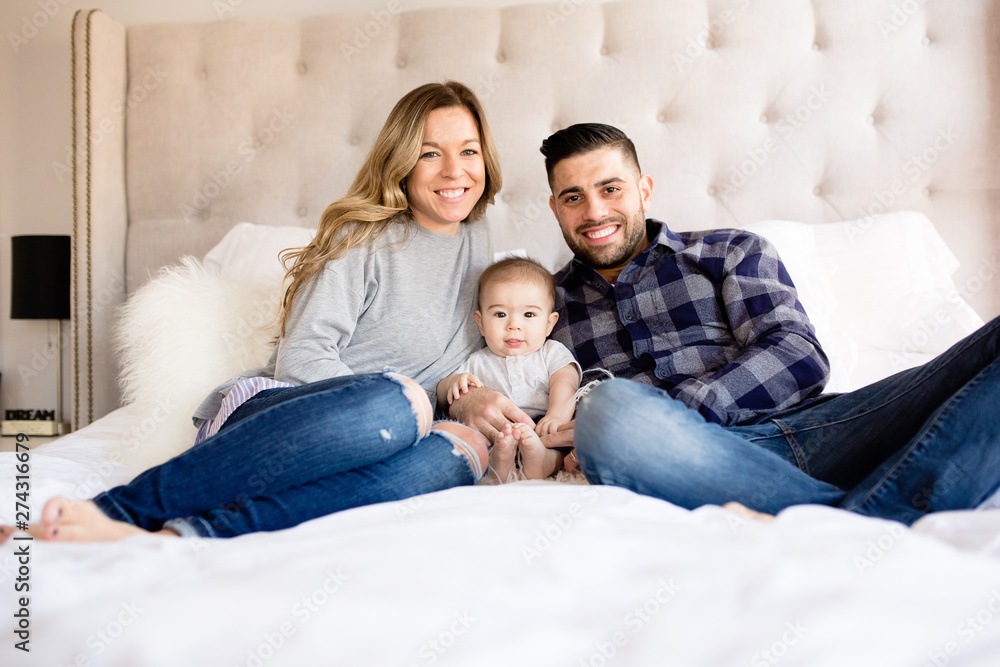 Mom dad and baby sitting on bed and smiling at camera Mom dad and baby sitting on bed and smiling at camera