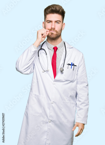 Young handsome doctor man wearing medical coat mouth and lips shut as zip with fingers. Secret and silent, taboo talking © Krakenimages.com