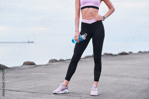 Young beautiful women in activewear and with bottle of water is posing for photographer at seaside.