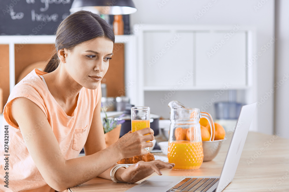 Smiling pretty woman looking at mobile phone and holding glass of orange juice while having breakfast in a kitchen