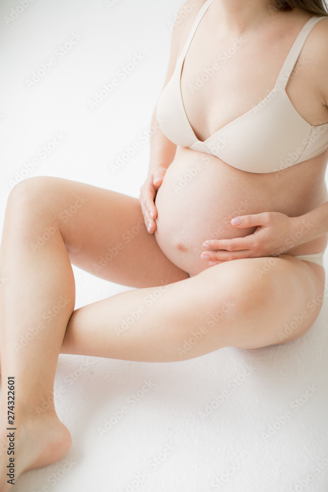 Happy young mother in anticipation of the birth of the baby. A pregnant girl is sitting clasping her bare belly. On a white background. Happy young mother in anticipation of the birth of the baby. A pregnant girl is sitting clasping her bare belly. On a white background.