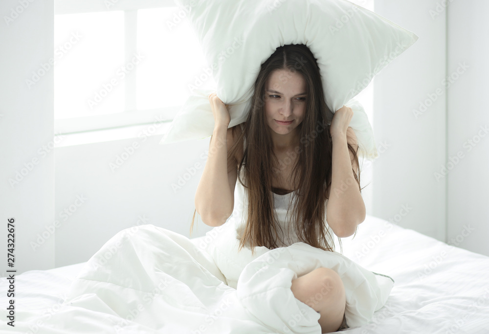Annoyed young woman covering ears with pillow, blocking sound, suffering from noise, lying in bed, problem with sleep Annoyed young woman covering ears with pillow, blocking sound, suffering from noise, lying in bed, problem with sleep