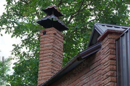 long brick brown chimney on the roof among the branches and green leaves