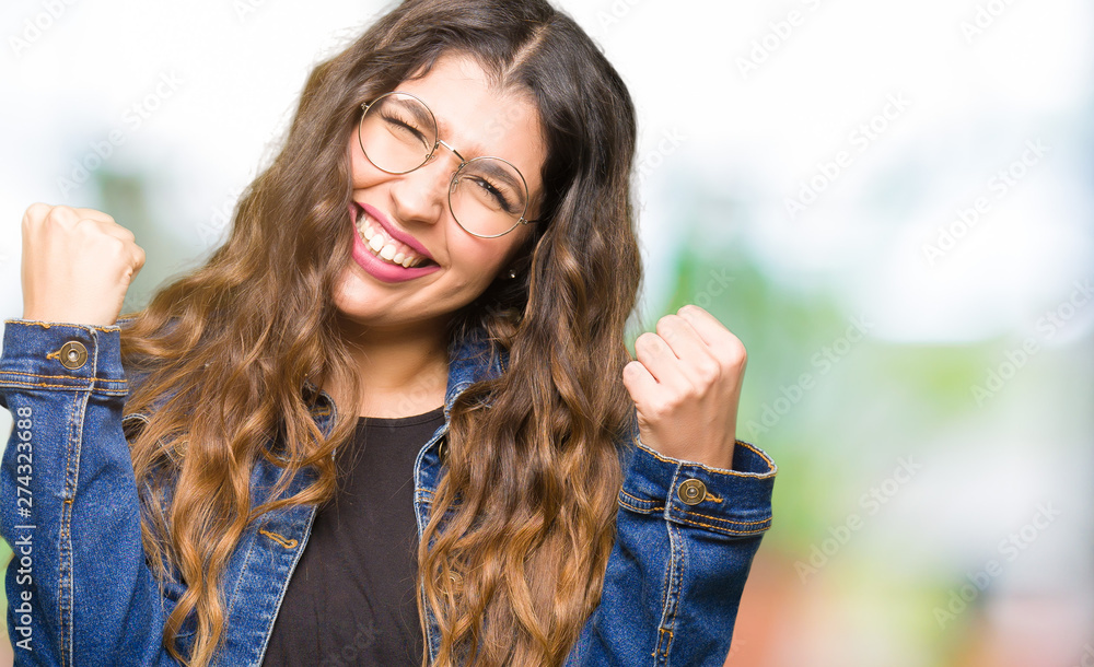 Young beautiful woman wearing glasses and denim jacket very happy and excited doing winner gesture with arms raised, smiling and screaming for success. Celebration concept.