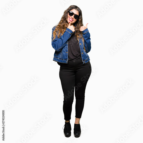 Young beautiful woman wearing sunglasses doing happy thumbs up gesture with hand. Approving expression looking at the camera with showing success.