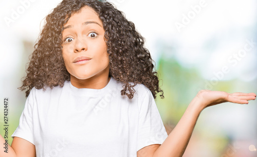 Young beautiful woman with curly hair wearing white t-shirt clueless and confused expression with arms and hands raised. Doubt concept. © Krakenimages.com