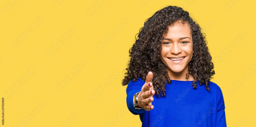 Young beautiful woman with curly hair wearing winter sweater smiling friendly offering handshake as greeting and welcoming. Successful business. Young beautiful woman with curly hair wearing winter sweater smiling friendly offering handshake as greeting and welcoming. Successful business.