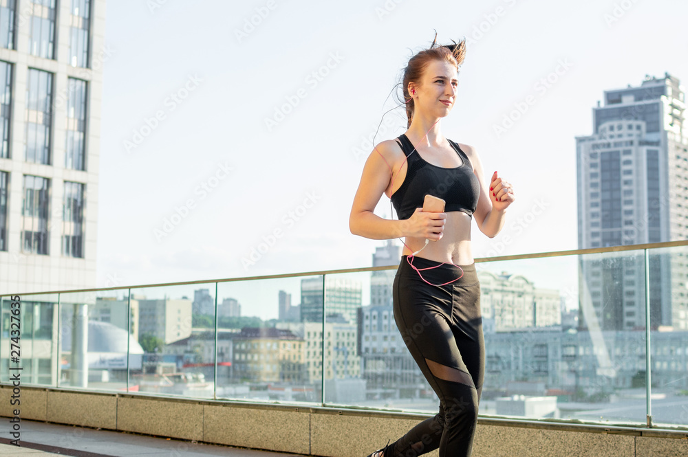 athletic girl runs to workout in the morning against the background of the city and listens to music, she smiles and trains