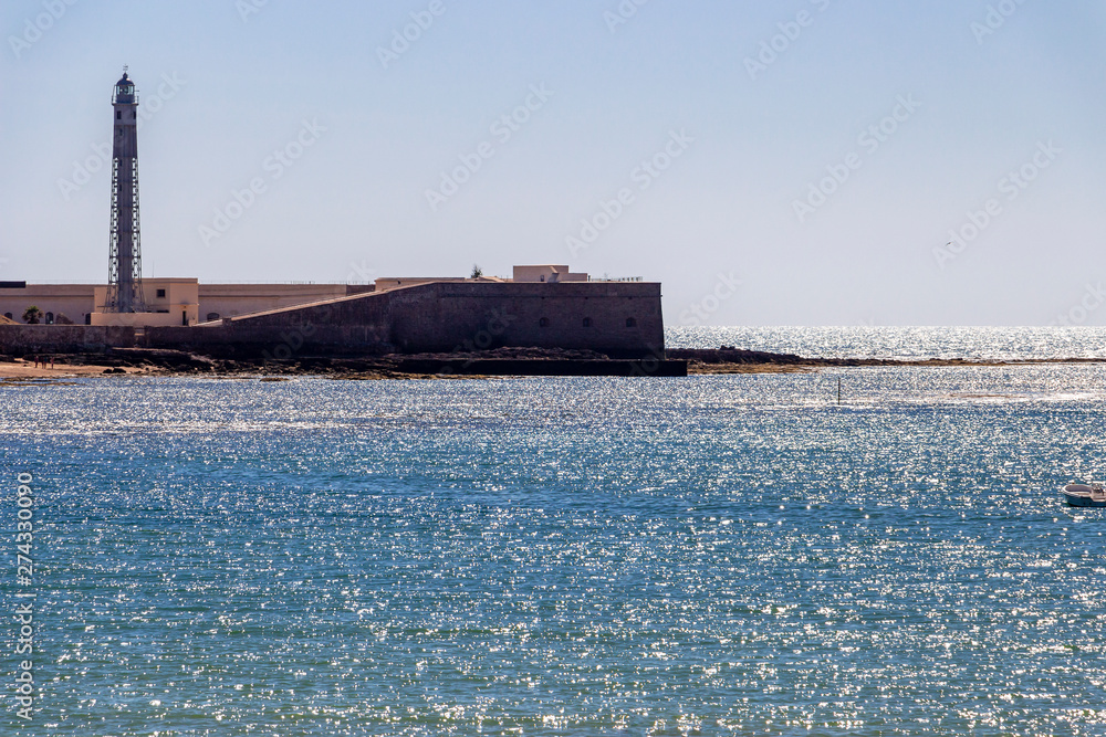 Fototapeta premium Shimmering Atlantic Ocean water surface with part of the Castle of San Sebastian and the lighthouse in Cadiz, Province of Cadiz, Andalusia, Spain seen from Playa la Caleta