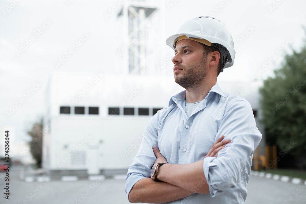 Young handsome male engineer in hardhat standing on the background of water or gas pipes. Concept of boiler station. Pipeline accessories closeup. Heating station manager doing his job outdoor. . Young handsome male engineer in hardhat standing on the background of water or gas pipes. Concept of boiler station. Pipeline accessories closeup. Heating station manager doing his job outdoor. .