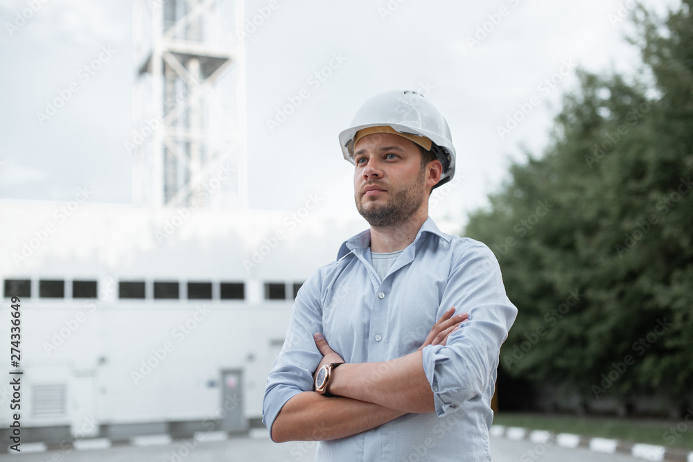 Young handsome male engineer in hardhat standing on the background of water or gas pipes. Concept of boiler station. Pipeline accessories closeup. Heating station manager doing his job outdoor. . Young handsome male engineer in hardhat standing on the background of water or gas pipes. Concept of boiler station. Pipeline accessories closeup. Heating station manager doing his job outdoor. .