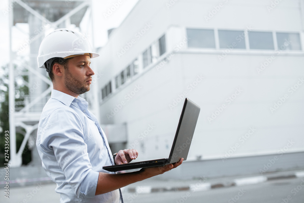 Young engineer in hardhat standing on the background of water or gas pipes station and using laptop. Heating station manager doing his job outdoor. Young engineer in hardhat standing on the background of water or gas pipes station and using laptop. Heating station manager doing his job outdoor.