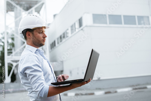 Young engineer in hardhat standing on the background of water or gas pipes station and using laptop. Heating station manager doing his job outdoor.