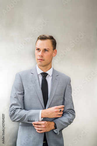 Portrait of Young European Businessman in New York City. Young Man wearing gray blazer, white shirt, black tie, hand touching cuff, standing against silver metal background, confidently looking up..