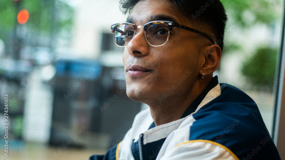 Chicago, IL/United States-June 12th 2019: Young attractive Middle eastern male model drinks his coffee from behind the window glass at a local Chicago shop. the student ponders about the day Chicago, IL/United States-June 12th 2019: Young attractive Middle eastern male model drinks his coffee from behind the window glass at a local Chicago shop. the student ponders about the day