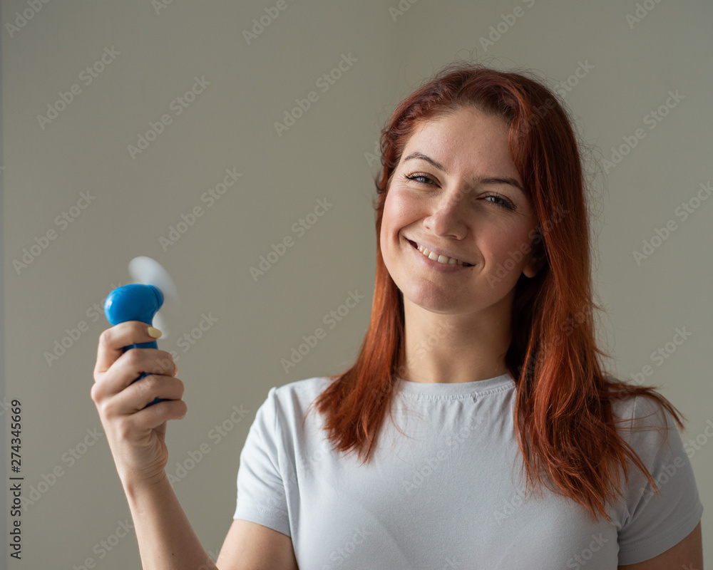 Portrait of a red-haired smiling woman with a pocket fan in her hands. Full face of a girl refreshing in the heat of a wind blow from a wireless electric device. Cool air flow from the air conditioner Portrait of a red-haired smiling woman with a pocket fan in her hands. Full face of a girl refreshing in the heat of a wind blow from a wireless electric device. Cool air flow from the air conditioner