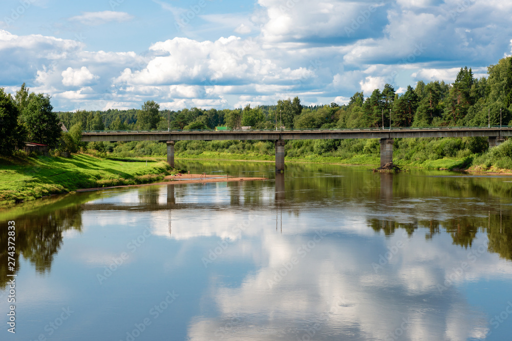River landscape. Road bridge across the river Msta and clouds reflected in the water on a summer day.