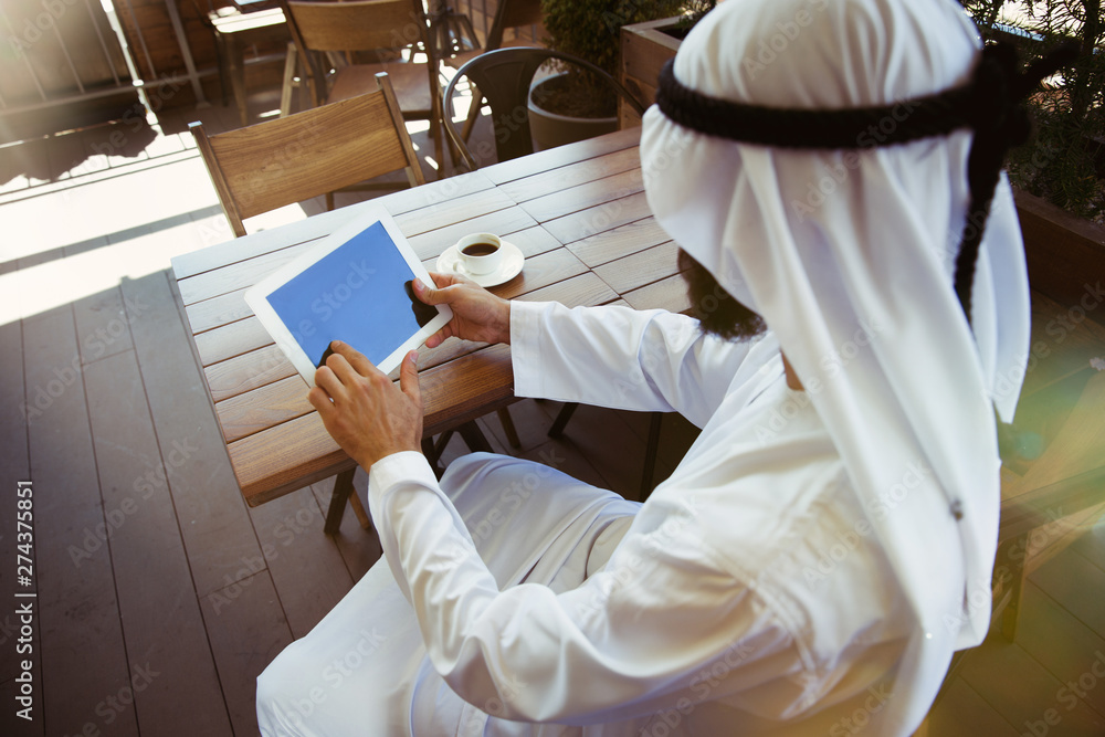 Arab saudi businessman working online with a laptop and tablet in a coffee shop or a cafe with an outdoor terrace in the background. Concept of business, finance, modern technologies, start up. Arab saudi businessman working online with a laptop and tablet in a coffee shop or a cafe with an outdoor terrace in the background. Concept of business, finance, modern technologies, start up.