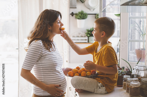 Pregnant mom with kid eating fruits together in the kitchen
