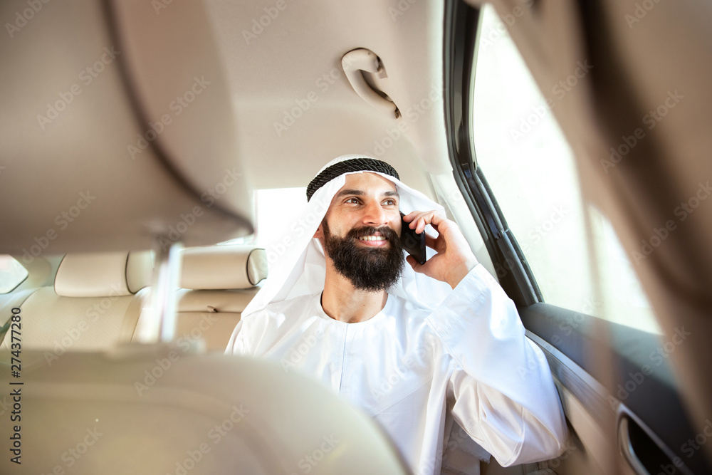 © master1305 - Arab saudi businessman working online with a smartphone, calling his clients or partners in his car. Male model as an enterpreneur. Concept of business, finance, modern technologies, start up, economy