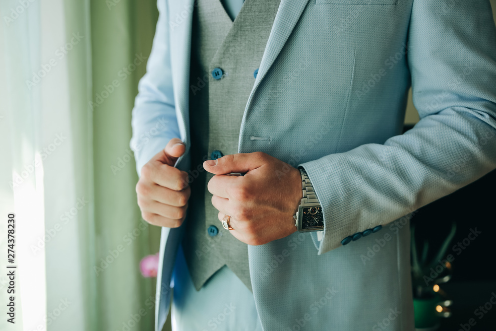 Groom in light blue suit is standing near the window. Man is wearing his jacket. Smart clothes.