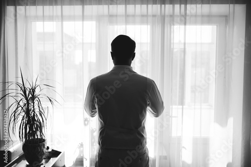 Groom in light grey suit is standing near the window. Man looking in front of him. Black and white photo.