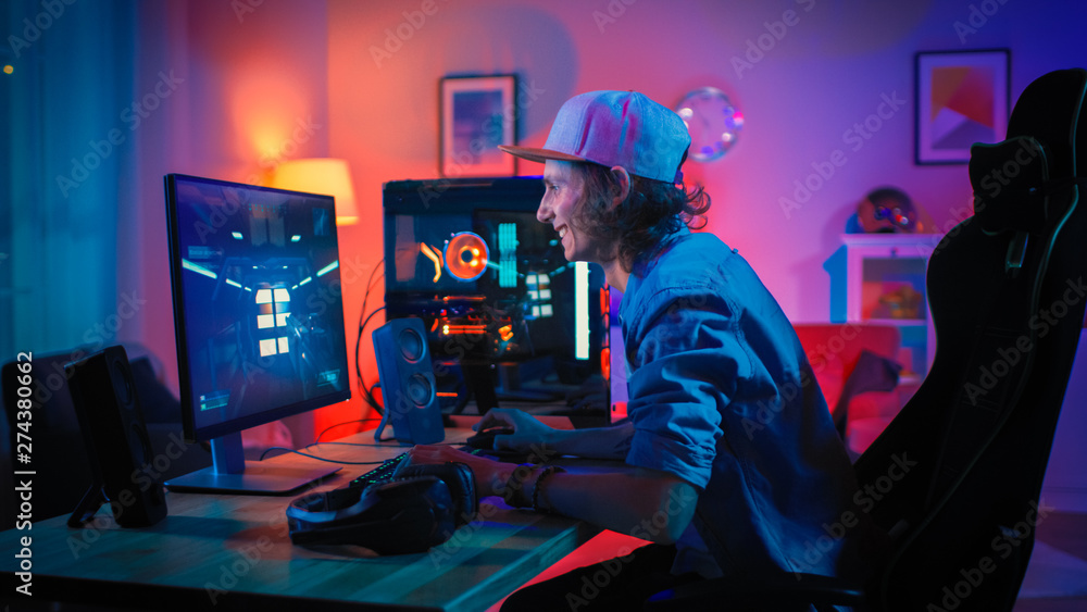Professional Gamer Playing First-Person Shooter Online Video Game on His Powerful Personal Computer with Colorful Neon Led Lights. Young Man is Wearing a Cap. Living Room Lit with Warm Red Lamps. - obrazy, fototapety, plakaty 