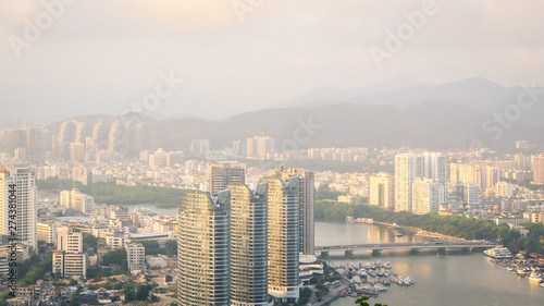 Fototapeta Naklejka Na Ścianę i Meble -  View of one of the districts of Sanya city. Visible are the skyscrapers and the public ferry terminal. Sunset, blur, haze. Hainan Island, China
