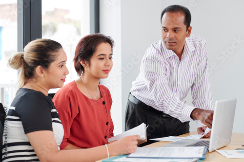 Businessman pointing at laptop computer and explaining online work to two businesswomen sitting at the table and looking at monitor