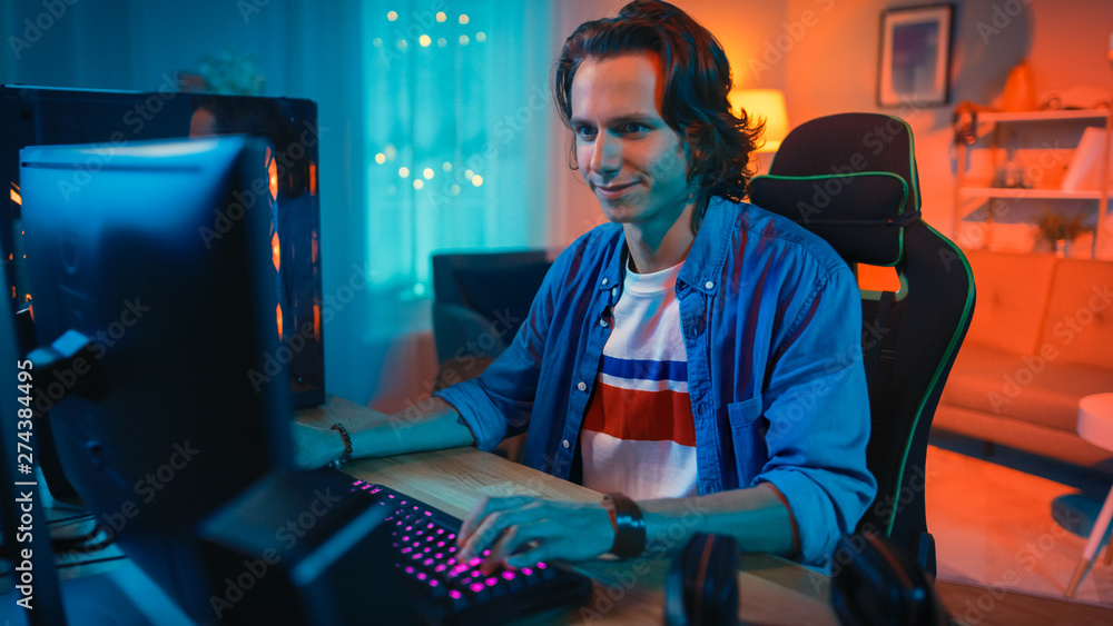 Excited Gamer Playing Online Video Game on His Personal Computer. Room and Personal Computer have Colorful Warm Neon Led Lights. Young Man has Long Hair and Handsome Smile. Cozy Evening at Home.