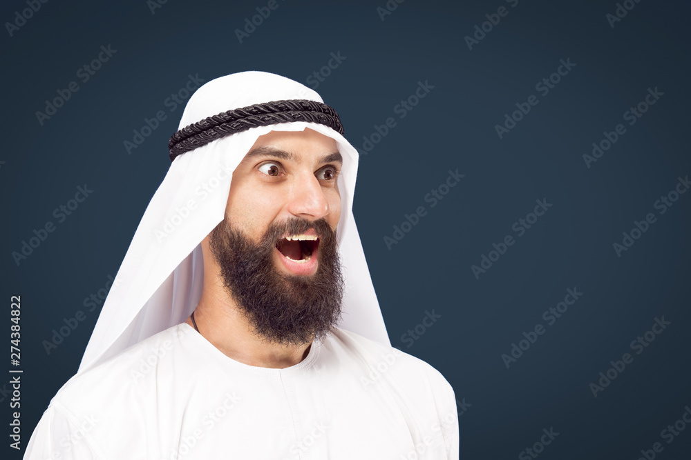 Half-length portrait of arabian saudi businessman on dark blue studio background. Young male model posing and smiling, looks happy. Concept of business, finance, facial expression, human emotions. Half-length portrait of arabian saudi businessman on dark blue studio background. Young male model posing and smiling, looks happy. Concept of business, finance, facial expression, human emotions.