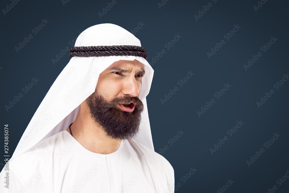 Half-length portrait of arabian saudi businessman on dark blue studio background. Young male model posing and looks serious or angry. Concept of business, finance, facial expression, human emotions. Half-length portrait of arabian saudi businessman on dark blue studio background. Young male model posing and looks serious or angry. Concept of business, finance, facial expression, human emotions.