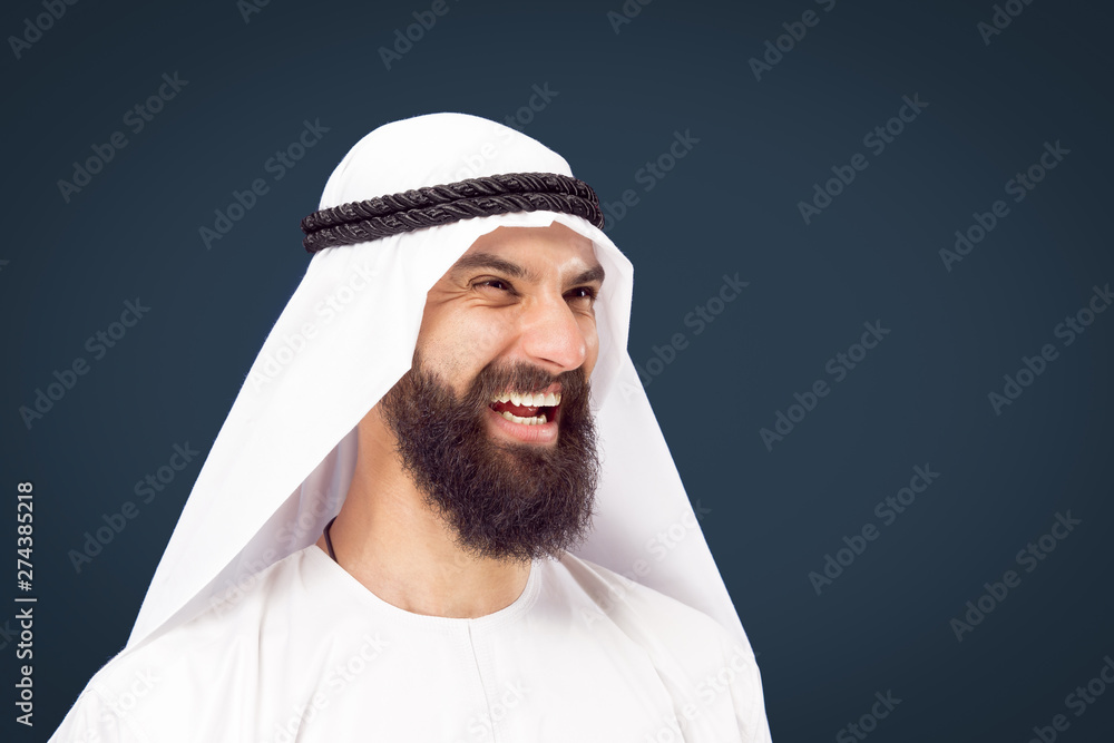 Half-length portrait of arabian saudi businessman on dark blue studio background. Young male model posing and smiling, looks happy. Concept of business, finance, facial expression, human emotions. Half-length portrait of arabian saudi businessman on dark blue studio background. Young male model posing and smiling, looks happy. Concept of business, finance, facial expression, human emotions.