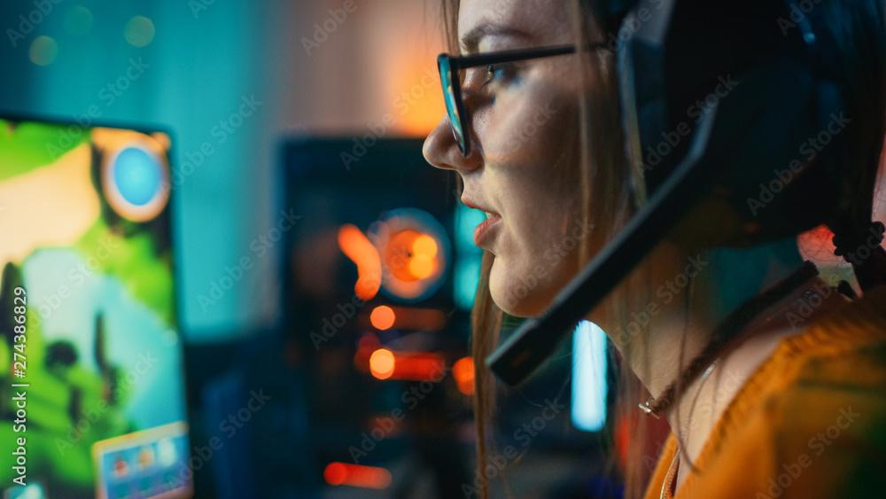 Close-up of an Excited Gamer Girl in Headset with a Mic Playing Online Strategy Video Game on Her Personal Computer. Room and PC have Colorful Warm Neon Led Lights. Cozy Evening at Home.