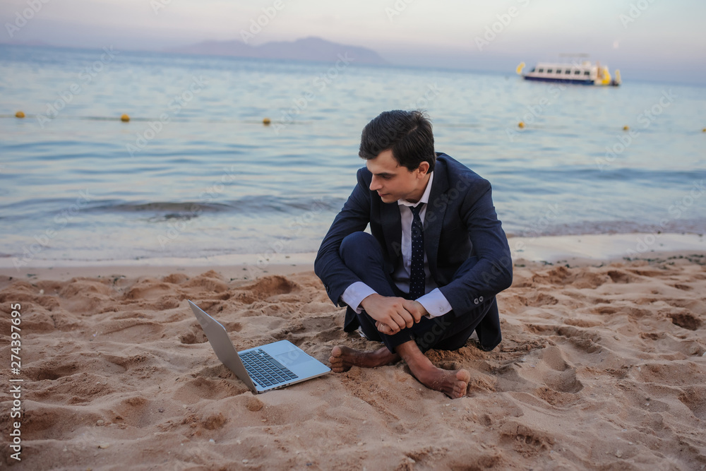 lateral view of a young man in suit with laptop sitting on the beach working on the seaside lateral view of a young man in suit with laptop sitting on the beach working on the seaside