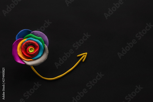 A flower made from plasticine with an arrow pointing up from it on black background, shot from above.