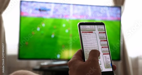 Against the background of the TV at home (at the bar), the guy (woman) orders different snacks, pizza and sweets scrolling through internet stores using mobile phones. Concept: Football, Sport,Order.