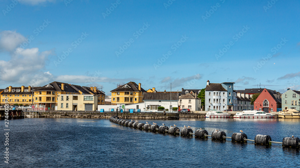 Obraz premium River Shannon with plastic buoys on the calm water of the river, cityscape with quaint houses in the town of Athlone in background, sunny day with a blue sky in County Westmeath, Ireland