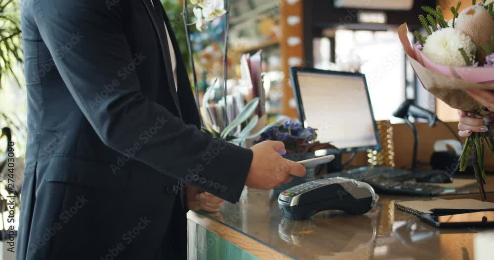 Man using smartphone to make payment in florist shop then taking bouquet purchasing gift with electronic money. People, devices and consumerism concept. Man using smartphone to make payment in florist shop then taking bouquet purchasing gift with electronic money. People, devices and consumerism concept.