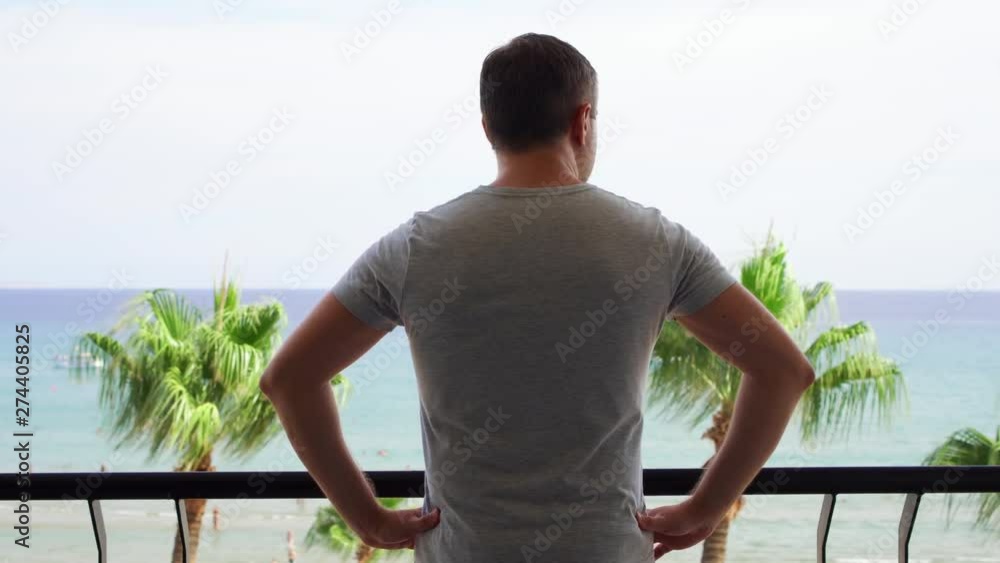 Young man going out onto balcony and enjoying view from terrace. Male on vacation looking at Mediterranean sea. Wind swaying leaves on palm trees Young man going out onto balcony and enjoying view from terrace. Male on vacation looking at Mediterranean sea. Wind swaying leaves on palm trees