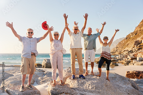 Portrait Of Senior Friends Standing On Rocks By Sea On Summer Group Vacation With Arms Outstretched