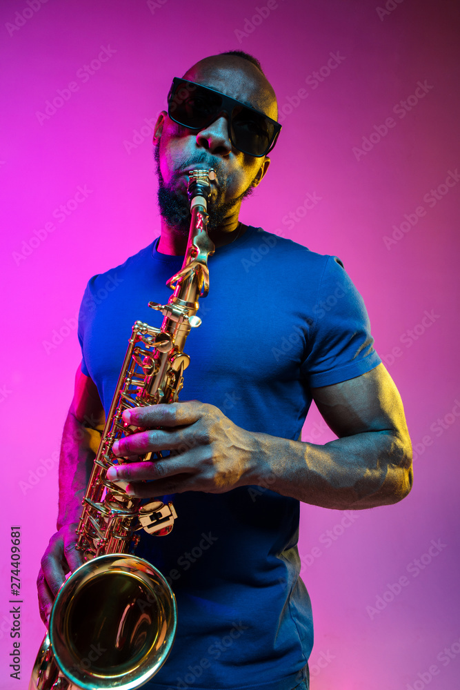 Fototapeta premium Young african-american jazz musician playing the saxophone on pink studio background in trendy neon light. Concept of music, hobby. Joyful attractive guy improvising. Colorful portrait of artist.