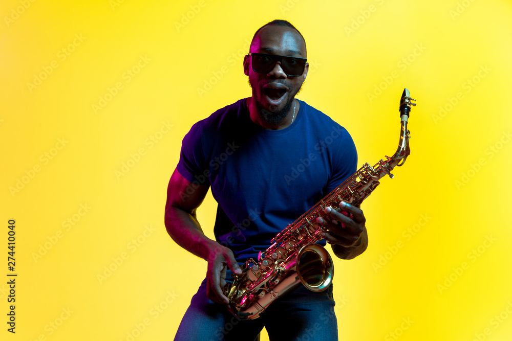 Naklejka premium Young african-american jazz musician playing the saxophone on yellow studio background in trendy neon light. Concept of music, hobby. Joyful attractive guy improvising. Colorful portrait of artist.