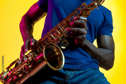 Photography Young african-american jazz musician playing the saxophone on yellow studio background in trendy neon light