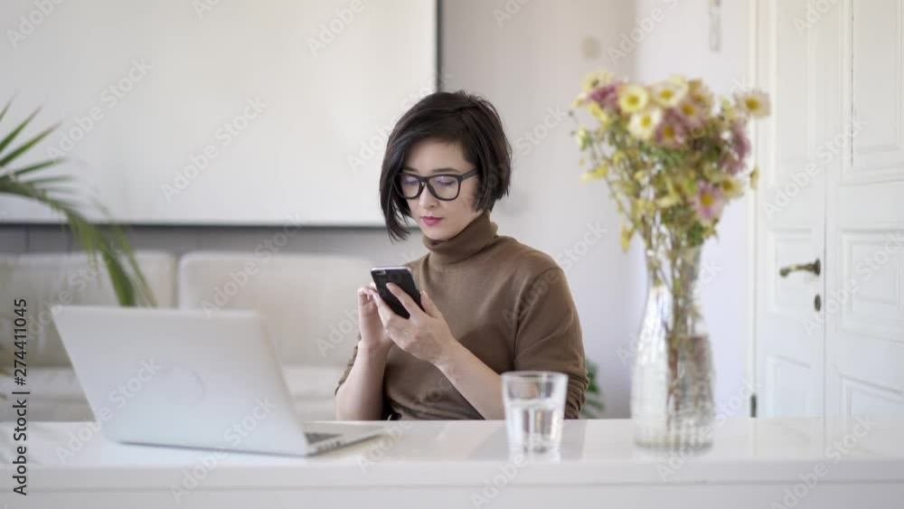Gimbal shot of occupied asian business woman in eyeglasses with short haircut scrolling the phone. Laptop, glass of water on table and a vase with flowers in white home office Gimbal shot of occupied asian business woman in eyeglasses with short haircut scrolling the phone. Laptop, glass of water on table and a vase with flowers in white home office